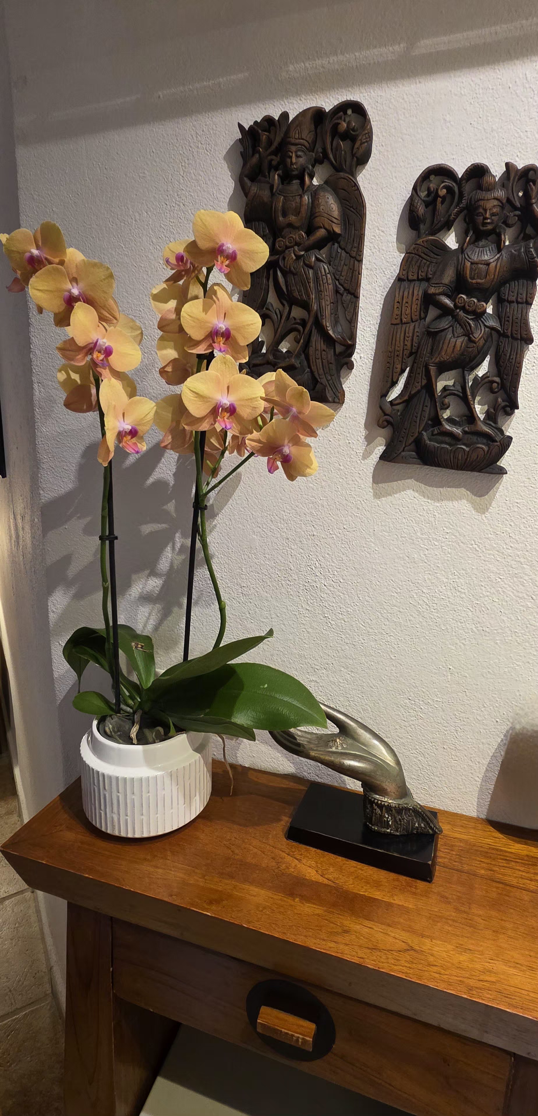 Yellow orchids in a white pot beside carved wooden figures on a console