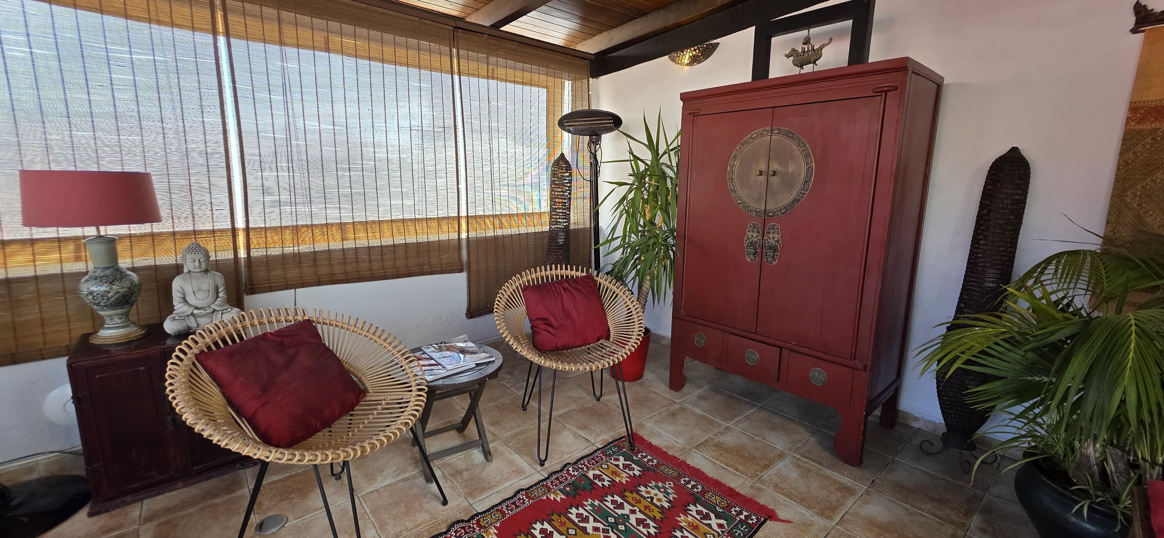 A guest room at La Villa Marbella with a red lacquered cabinet, wicker armchair, and bamboo blinds at the window